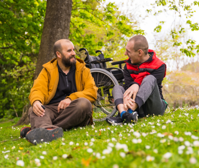 Beeld van twee mannen die in het gras zitten, rolstoel op de achtergrond. Ze lachen naar elkaar. Een van hen is de mantelzorger en de ander is de zorgbehoevende.
